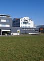 Commercial building with sign 'Migros', grey exterior, multiple windows, open garage door, cars parked in front, spacious lawn area