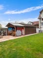 Two-story residential building with a tiled roof, surrounded by a grassy yard and a paved patio area. The yard features a wooden shed or storage structure, a barbecue grill, and other outdoor furniture. There is also a garage or carport visible on the pro