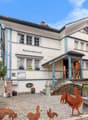 Two-story building with a traditional German architectural style, featuring a restaurant called 'Speiserestaurant' on the ground floor. The building has a tiled roof, large windows, and decorative elements. There are some rustic metal sculptures of animal