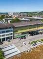 Large office building with yellow and green exteriors, numerous windows, parking lot with cars parked, solar panels on the roof, surrounded by greenery and other buildings