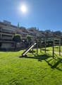 Playground with a slide and a bench, surrounded by green grass and trees, with a walkway leading to the building