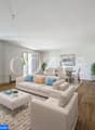 Spacious open-plan living room with large windows, hardwood flooring, beige sofa set, coffee table, and dining area with table and chairs. The room has a bright and airy feel with natural light and potted plants.