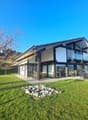House with modern design, large windows, terrace with seating area, lush lawn, patio with fireplace, surrounded by greenery.
