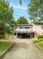 Two-story house with a pink exterior, surrounded by lush greenery and trees. The house has a balcony on the upper level and a garage with a parking space underneath. The property has a paved walkway leading to the entrance and a well-maintained lawn.