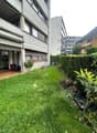 Building with tiled entrance, open garage, small garden area, balcony with potted plants, hedge, grass lawn, trash bins, and a tool cart.