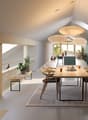 The image shows an open concept living, dining, and kitchen area with a modern and minimalist design. The space features high ceilings, large windows that provide natural light, and a wooden dining table with chairs. The kitchen area has green cabinets an