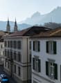 This image shows a picturesque street in a European city, with traditional buildings featuring white facades, black shutters, and red tile roofs. In the background, snow-capped mountains can be seen, creating a stunning natural backdrop. The street is lin