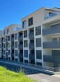 Modern apartment building, multiple floors, balconies, glass railings, white exterior, green grass, street view