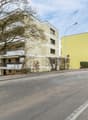 Large multi-story building with balconies, beige exterior, modern design, surrounded by trees and bushes, clear sky