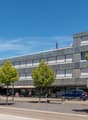 Modern building with multiple floors, gray exterior, large windows, parking lot, trees, cars parked in front