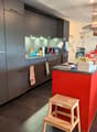 Modern kitchen, gray cabinets, red island counter, wooden step stool, wooden cabinets on the right side, stainless steel range hood, tiled floor.