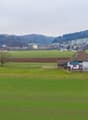 The image shows a rural countryside landscape with rolling green hills, a small village in the distance, and a farmhouse with a balcony or terrace in the foreground. The scene has a peaceful, pastoral feel with fields, fences, and a few trees dotting the