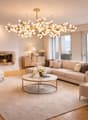 chandelier, white sofa, coffee table, fireplace, large windows, view, side tables, two armchairs, white armchair with cushion, beige rug, two floor lamps, two floor vases