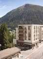 A modern apartment building with several balconies and windows, located on a slope with a view of the mountains.