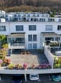 White multi-story building with multiple balconies, plants, flowers, cars parked in the ground floor parking, tables and chairs outside