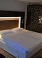 Modern bedroom with a large bed, white bedding, and a decorative black and white patterned wall panel. The room also features recessed lighting in the headboard and a fireplace or heating unit on the wall.