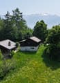 Two-story wooden cabin surrounded by lush green trees and vegetation, with a view of snow-capped mountains in the background.