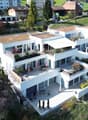Modern white apartment building, 3 stories, multiple balconies, terrace area with umbrellas, garden areas, view of the surrounding countryside.