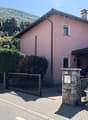 Two-story pink house with a tiled roof, surrounded by trees and greenery. The house has a garage and a stone wall with a gate in the front. There are potted plants and other decorative elements around the entrance.