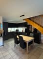 Dining area with a wooden table and black chairs, wooden staircase, kitchen with black cabinets, round mirror, and a wooden door.
