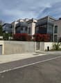 Modern apartment building with glass balconies, concrete wall, black railings, red bushes, and a concrete driveway.