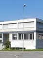 A modern building with many windows, possibly an office or industrial building, cars parked in front, white walls, a van parked in front