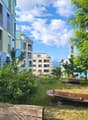 A garden with a grassy area, a circular bench, and a wooden log bench, surrounded by trees and bushes, with a view of the buildings in the background.