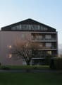 Apartment building, multiple floors, brick walls, black roofing, several balconies, large tree in front, green lawn