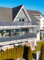 Two-story house, gray exterior, modern design, solar panels on roof, large glass terrace, paved front yard, hedges, nearby road.