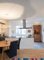 The image shows an open concept living space with a kitchen, dining area, and living room. The kitchen features a black range hood and built-in appliances. The dining area has a round wooden table with black chairs. The living room has a wooden cabinet an