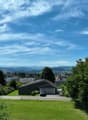 The image shows a scenic view of a residential area surrounded by lush greenery and mountains in the background. The sky is clear with wispy clouds, creating a picturesque setting. The houses in the foreground have a mix of architectural styles and are ne