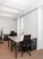 Spacious office space with large windows, hardwood floors, and modern black desks and chairs. The room has ample natural light and a view of the surrounding buildings outside.