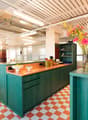 Open concept office kitchen with colorful, modern design. Features a large central island with wooden countertop, green cabinets, and a dishwasher. Bright lighting, checkered tile flooring, and decorative floral arrangement add to the vibrant atmosphere.