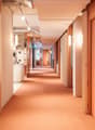 The hallway has wooden slat walls, a peach colored floor, and bright white lighting on the ceiling. At the end of the hallway is a kitchen area with wooden shelves, white tiles, and an island.