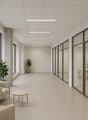 The image shows a modern, minimalist office space with a long hallway. The hallway has large windows along one side, providing natural light. The walls are painted in a neutral, light color, and the ceiling is covered with a grid of recessed lighting pane
