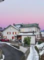 2 story house, white exterior, red shutters, snow on the roof, red car parked in front, pathway leading to the entrance, small garden with plants, fence, and a small wooden structure