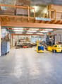 Empty warehouse with a large open area, wooden mezzanine with storage boxes, a few stacks of wooden pallets with boxes, two forklifts, machinery, metal barrels, and containers