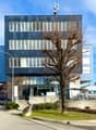 A multi-story office building with a modern glass and steel facade. The building has several floors and windows, and there is a tree in front of the entrance. The building appears to be located in an urban area with other buildings and vehicles visible in