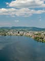 Aerial view of a scenic lakeside town surrounded by mountains and hills, with sailboats on the calm blue waters of the lake and a partly cloudy sky overhead.