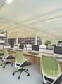 Modern office space with rows of ergonomic chairs, wooden desks, multiple monitors, keyboard and mouse, blue storage racks with boxes, artificial plants and ceiling lights.