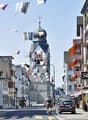 City street with buildings, church, flags, trees, cars, bicycles, street lights, signs, and potted plants.