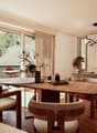 Spacious living room with large windows overlooking a balcony and outdoor greenery. The room features a wooden dining table with circular seating, a comfortable sofa, and a modern chandelier lighting fixture. The overall decor has a warm, natural aestheti