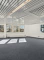 A large empty room with carpeted floor, multiple large windows, metal ceiling panels, recessed lighting, and no furnishings