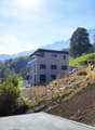 3-story modern residential building on a hillside, surrounded by trees and mountains. The building has a raised ground floor and is under construction, with construction equipment and materials visible on the site.