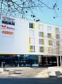 Modern white building with yellow accents, large glass windows, multiple floors, entrance with glass doors, signage for 'REX MAX' and 'COOP', concrete planters, street lights, and a tree in front.