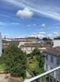 The image shows a panoramic view of a city skyline. The scene is dominated by a blue sky with fluffy white clouds. The cityscape is composed of a mix of residential and commercial buildings, with some greenery visible in the foreground. The buildings have