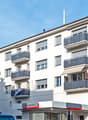 The image shows a multi-story apartment building with several balconies visible on the facade. The building has a modern, clean design with white walls and gray balcony railings. There is a commercial space on the ground floor, with the sign 'Auto Gambino