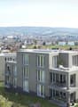 Multi-story apartment building with modern, gray exterior and large windows. The building has balconies on some units. The surrounding area includes a hilly, forested landscape with a small town or village in the distance.