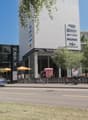 Building with vertical gardens, white walls, multiple signs indicating businesses. Umbrellas, bikes, and cars visible on the street. Trees line the sidewalk, and there are parked cars and bikes on the street.