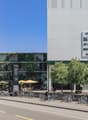 Multi-story building, vertical garden, parking area, street view, outdoor seating area, bikes parked on the side, car driving, trees and greenery, multiple shops on the ground floor, white walls and signs.